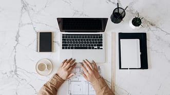 pexels-photo-6894103-6894103 Top view of a stylish home office desk with a laptop, planner, and coffee cup, showing hands on a blueprint.